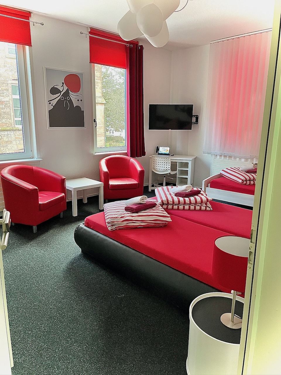 Dreibettzimmer mit rot bezogenen Betten, roten Sesseln und Flachbild-TV