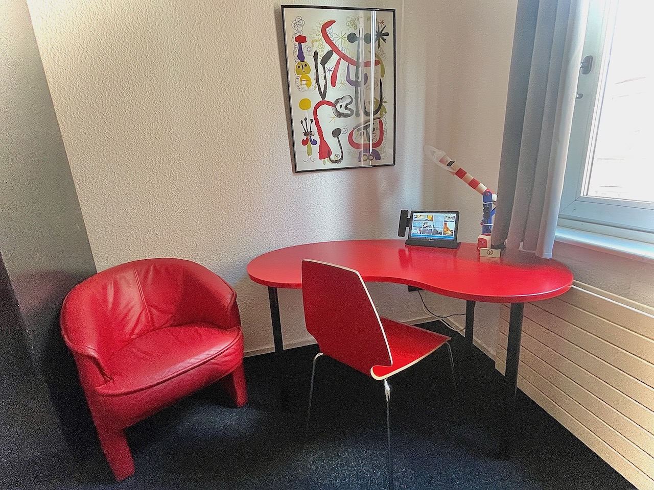 Einzelzimmer mit rotem Schreibtisch, rotem Stuhl und Miró-Druck