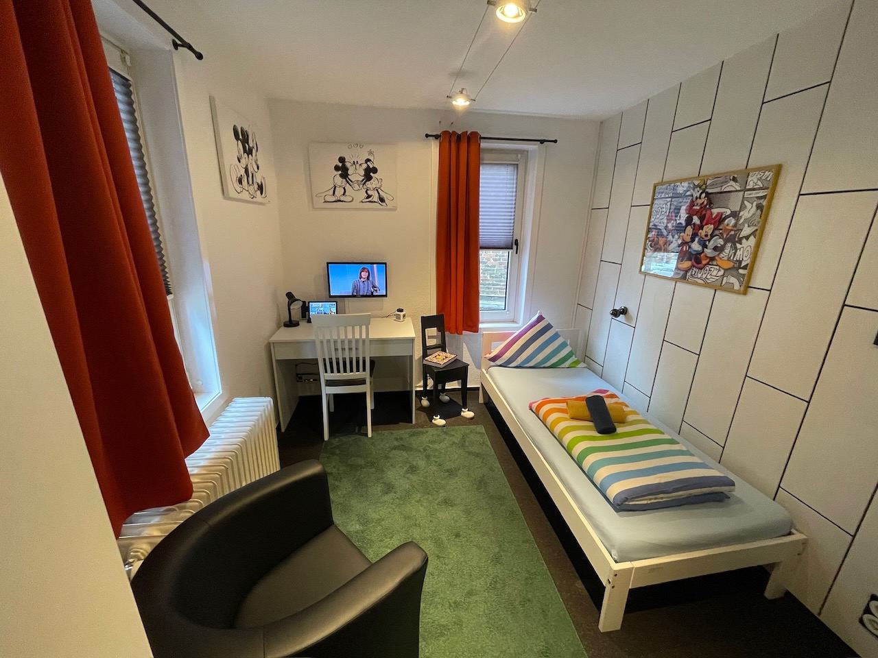 Einzelzimmer mit roten Vorhängen, Schreibtisch, Sessel und Bett