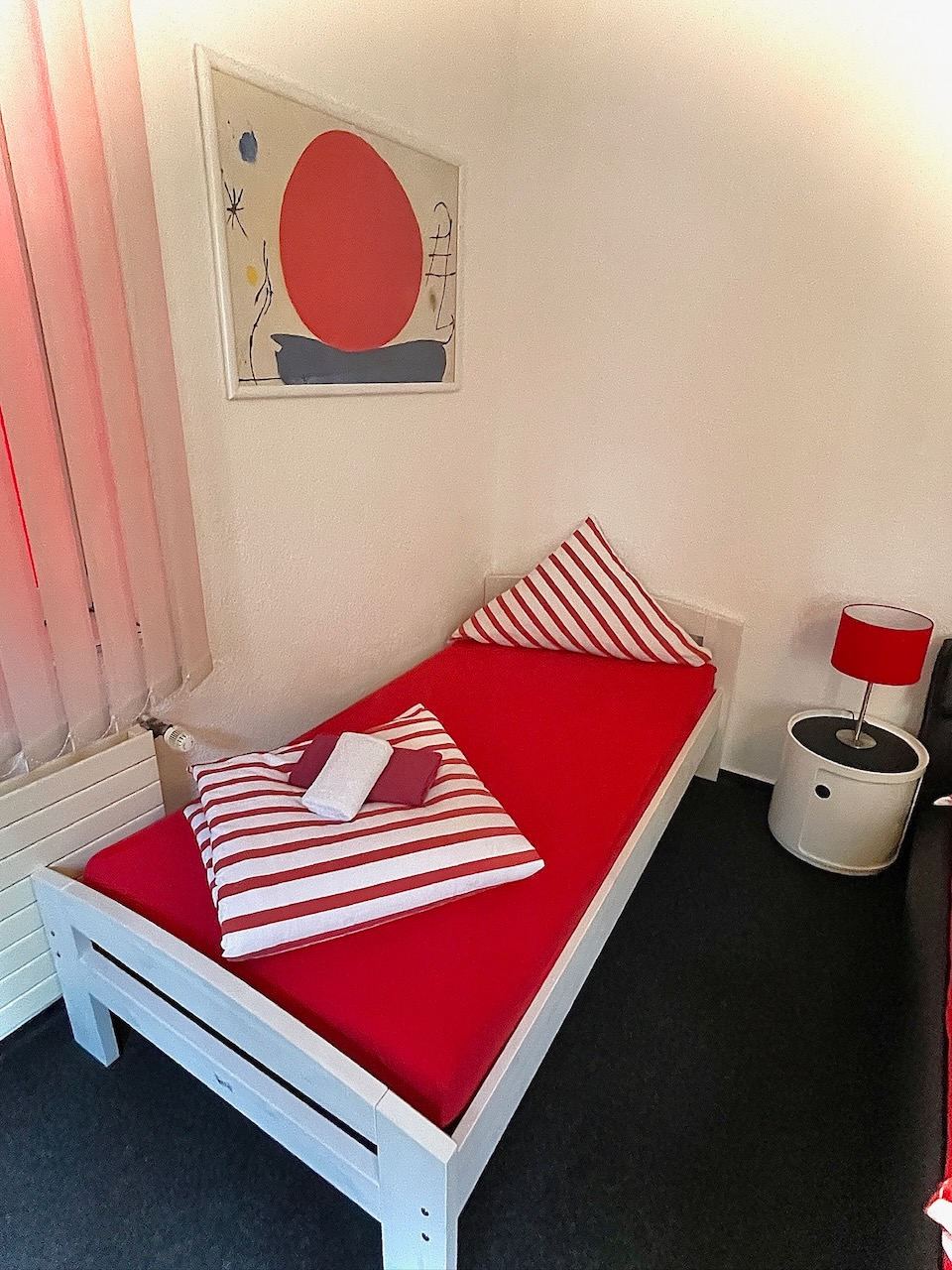 Einzelzimmer mit rot bezogenem Bett und Grafik über dem Kopfende