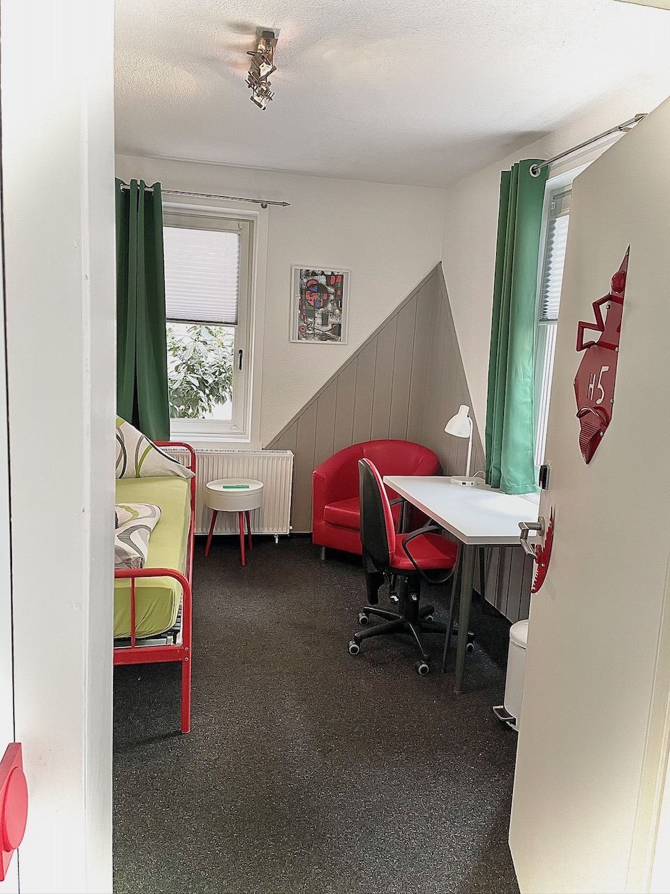 Einzelzimmer mit grünen Vorhängen, rotem Sessel und Schreibtisch
